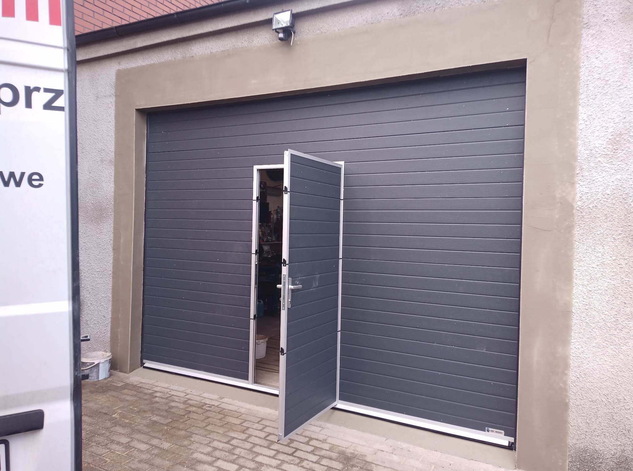 Szara brama garażowa segmentowa z drzwiami przejściowymi uchylonymi do wewnątrz, widoczny fragment elewacji budynku i kostki brukowej podjazdu.