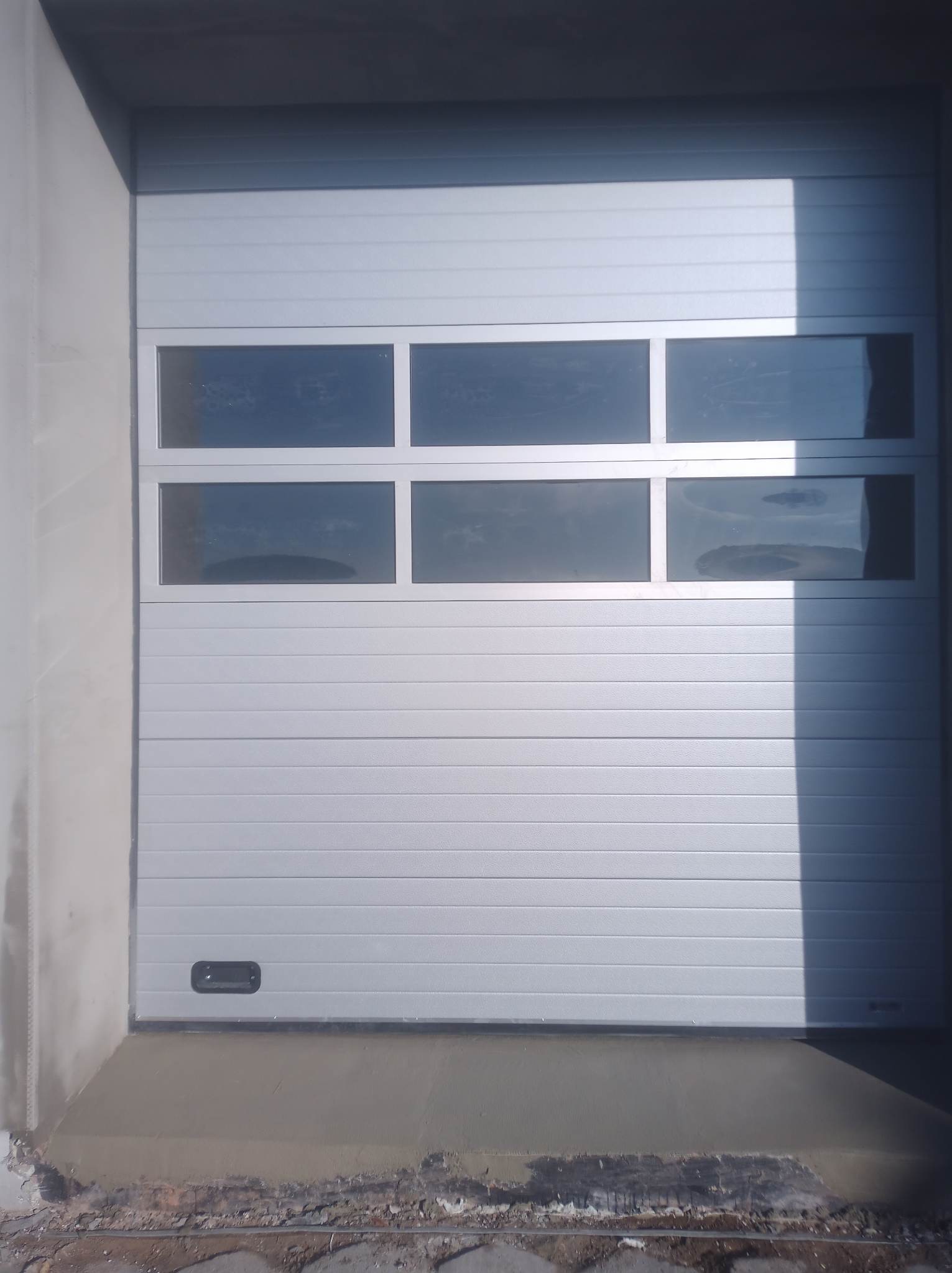 Zamknięta, segmentowa brama garażowa w kolorze jasnoszarym z poziomymi panelami i rzędem prostokątnych okienek u góry, zamontowana w surowym otworze garażowym z widocznymi śladami świeżego betonu.