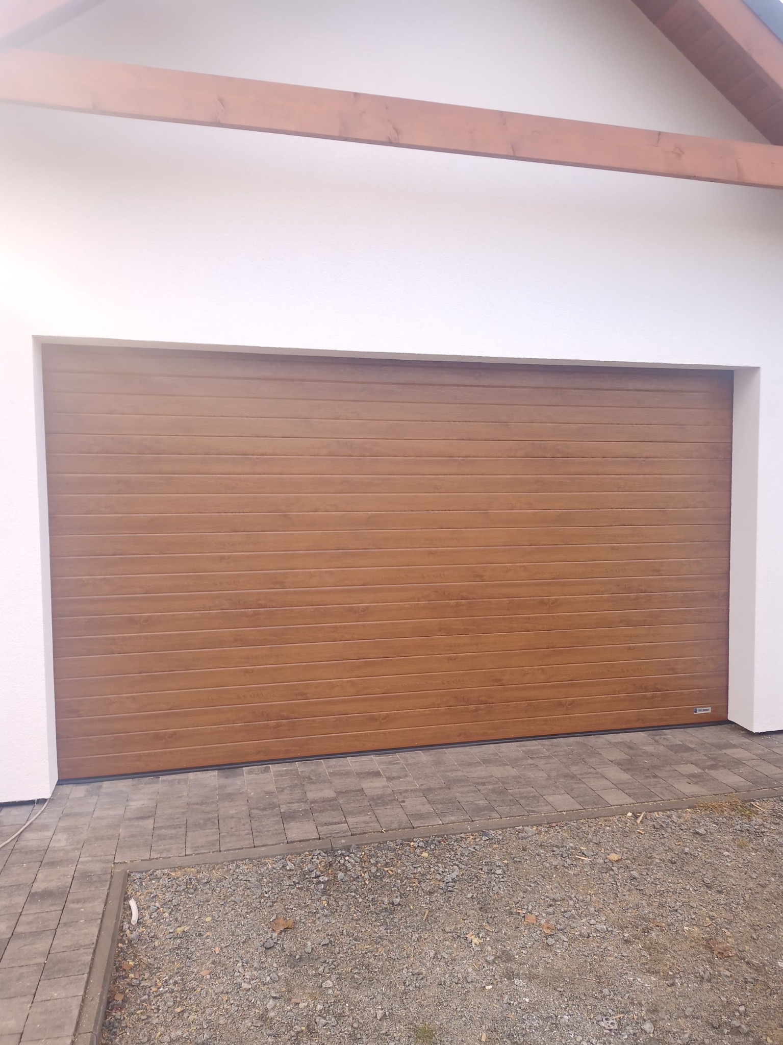 Zewnętrzna brama garażowa segmentowa w odcieniach brązu, z poziomymi przetłoczeniami imitującymi drewno, zamontowana w białej elewacji budynku z widoczną drewnianą belką stropową i brukowanym...