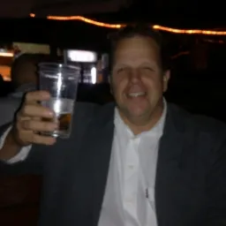 Uśmiechnięty mężczyzna w szarej marynarce i białej koszuli, trzymający plastikowy kubek z jasnym napojem, prawdopodobnie piwem, w barze z dekoracyjnym oświetleniem w tle.