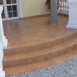 Brązowe płytki ceramiczne na schodach i tarasie domu z kolumną, widoczne kwiaty w oknach i kostka brukowa na zewnątrz.