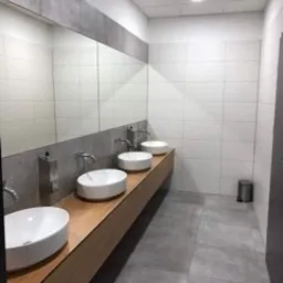 Nowoczesna łazienka publiczna z trzema białymi umywalkami nablatowymi na drewnianym blacie, duże lustro z szarą obwódką, szare płytki podłogowe i białe płytki ścienne z delikatnym wzorem, metalowy...