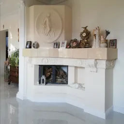 Klasyczny kominek z beżowego kamienia, zdobiony płaskorzeźbą i dekoracjami, z paleniskiem wypełnionym drewnem, ustawiony w przestronnym wnętrzu z kolumną.