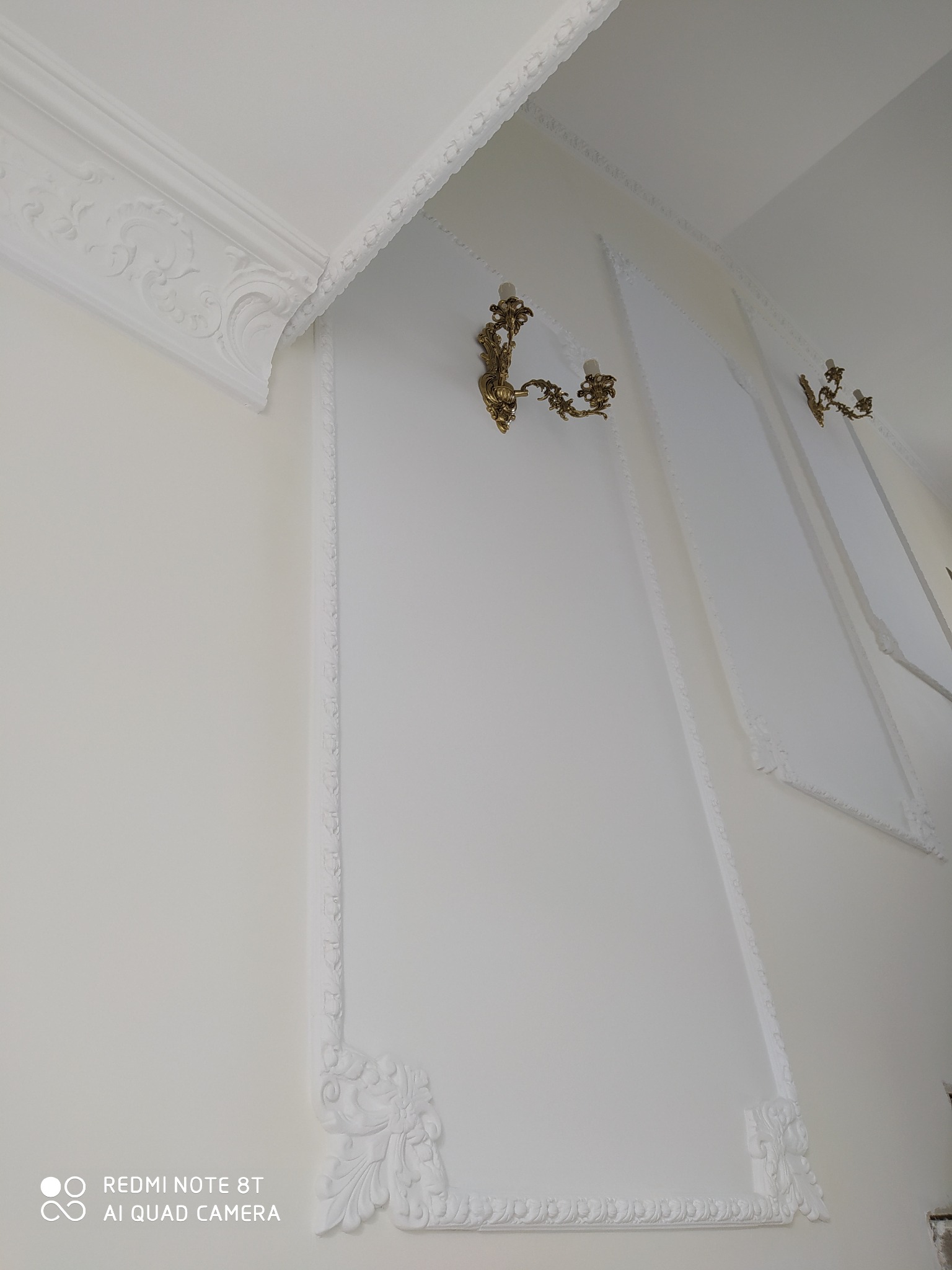 Białe ściany z dekoracyjnymi listwami i ramami, oświetlone złotymi kinkietami w stylu retro.