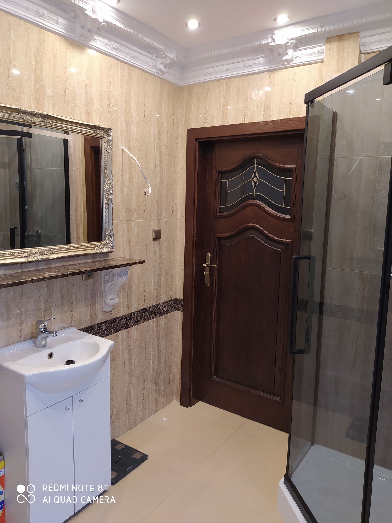 Elegancka łazienka z beżowymi płytkami, białym umywalką z szafką, lustrem w ozdobnej ramie, drewnianymi drzwiami z szybą i kabiną prysznicową z czarnymi profilami.