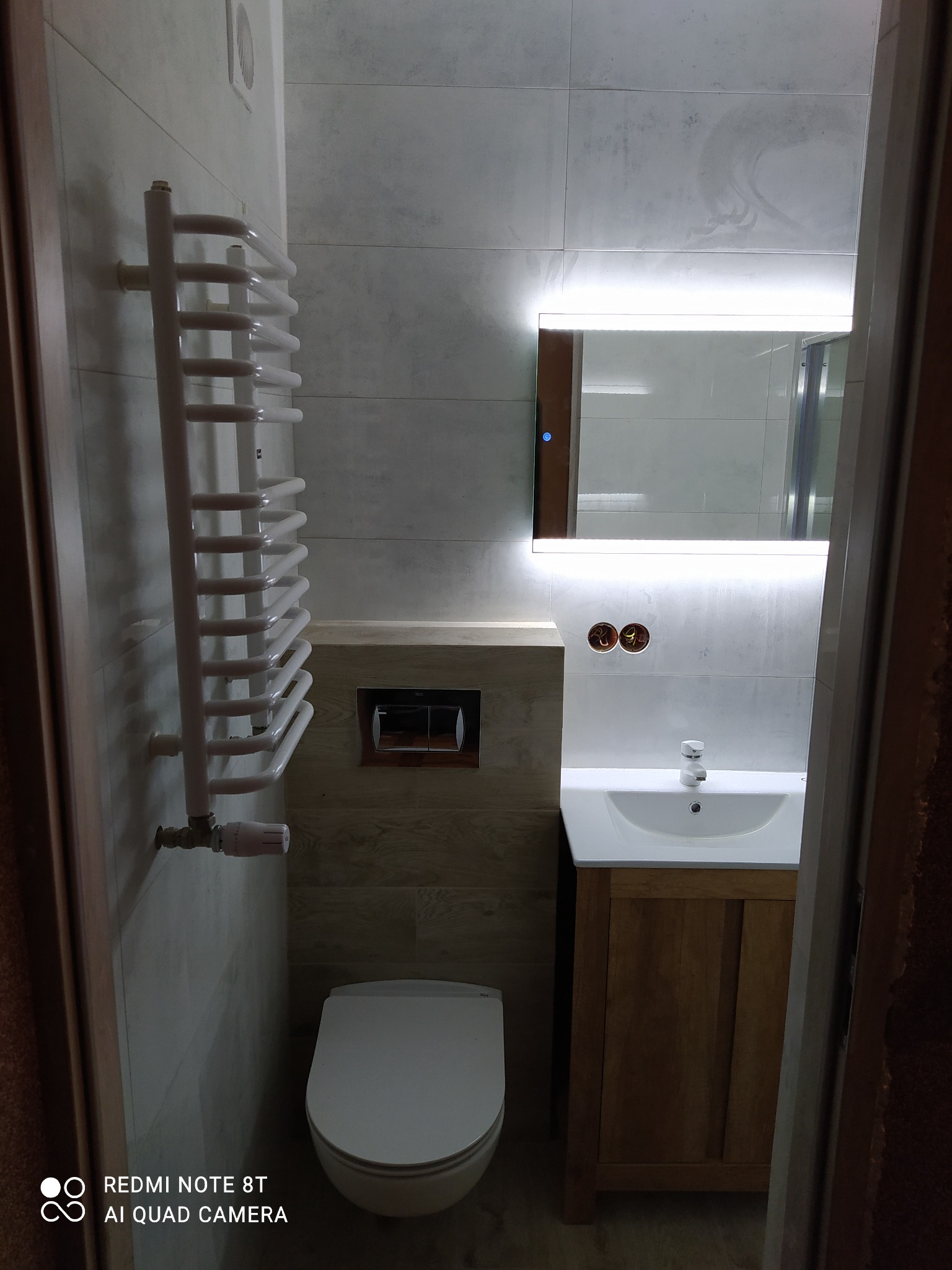 Nowoczesna łazienka po remoncie z podświetlanym lustrem, grzejnikiem drabinkowym i drewnianą szafką pod umywalką, widziana z perspektywy wejścia.