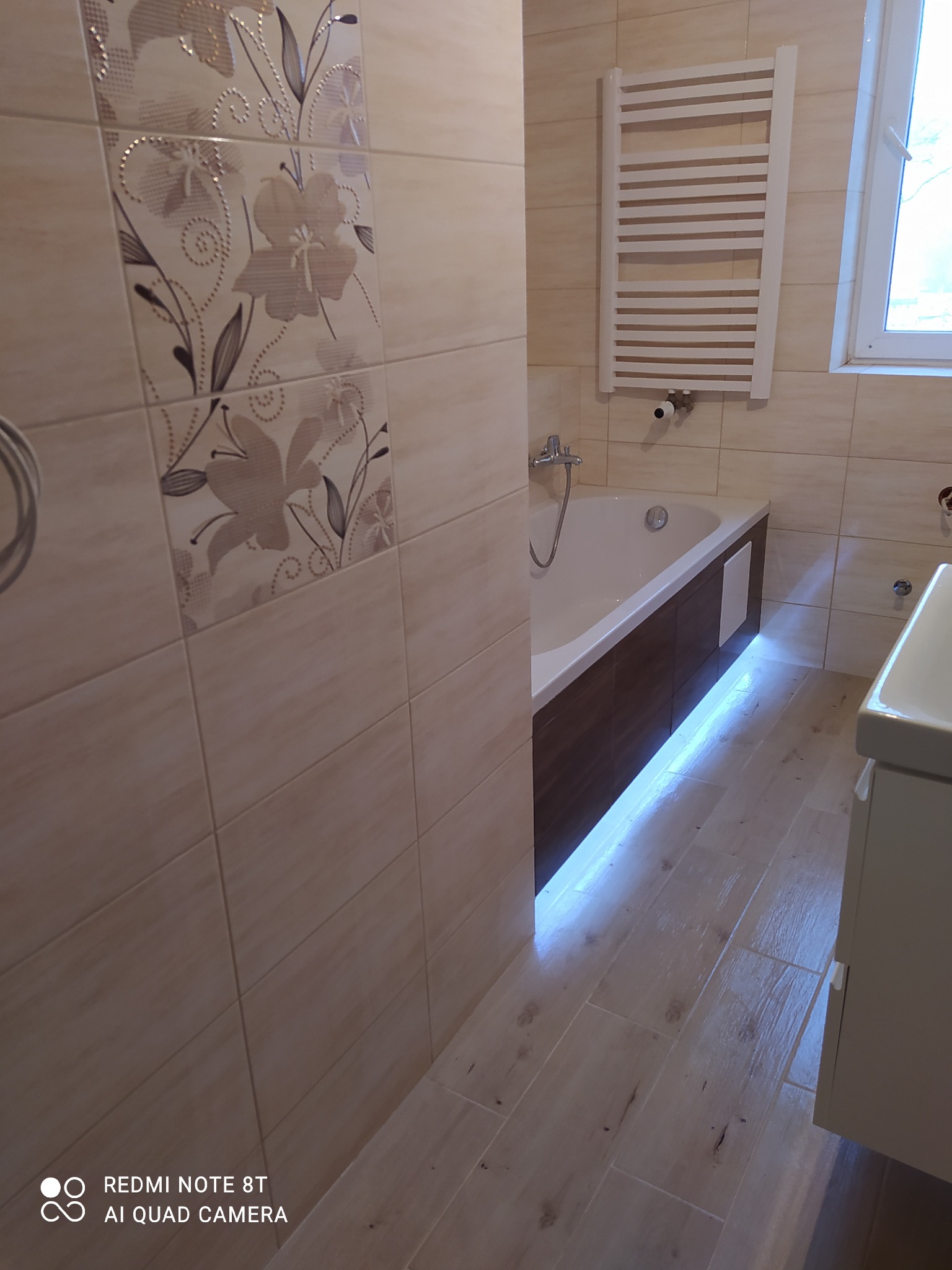 Wykończona łazienka z beżowymi płytkami, dekoracyjnym motywem kwiatowym na ścianie, podświetloną wanną z brązową obudową i grzejnikiem drabinkowym.