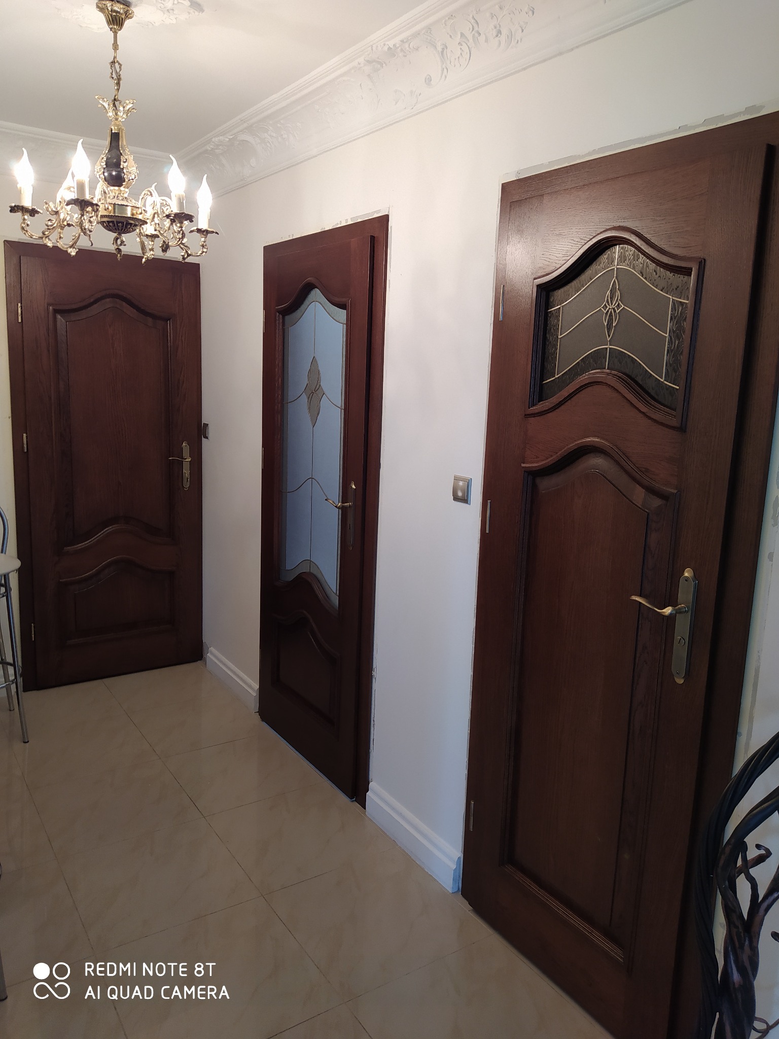 Korytarz z trzema ciemnymi, drewnianymi drzwiami o różnym wzornictwie, widoczna ozdobna listwa sufitowa i żyrandol.