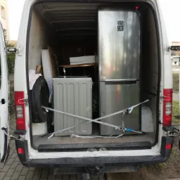 Wnętrze białego busa z otwartymi drzwiami, załadowane sprzętem AGD: srebrną lodówką Samsung i pralką, zabezpieczonymi pasami transportowymi, widoczne również opona i inne przedmioty.