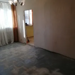 Pokój w trakcie demontażu podłogi z płytek, widoczne ślady po kleju i częściowo usunięte płytki, ściany w stanie surowym, w tle widoczne okno z firanką.