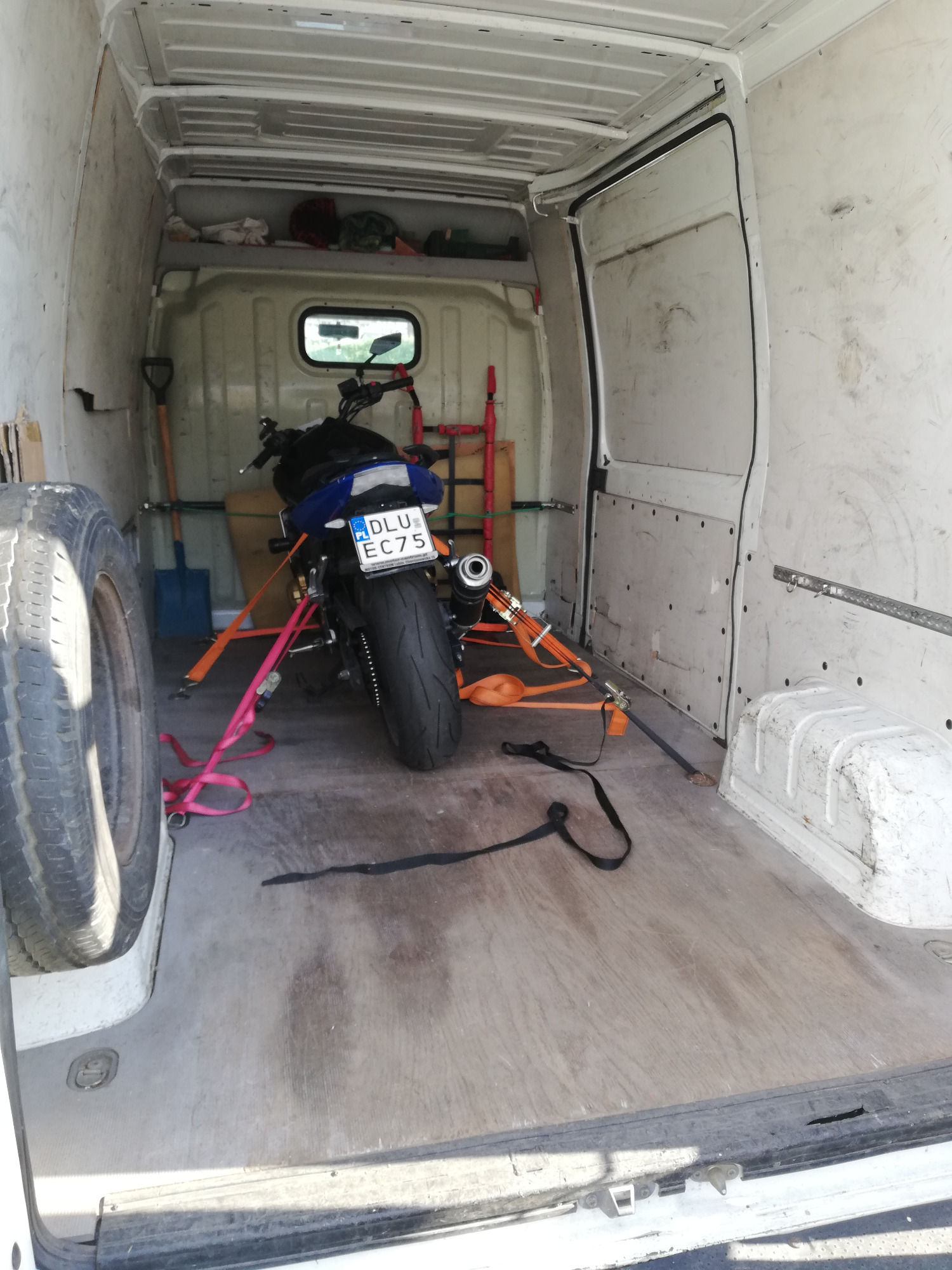 Wnętrze białego busa z zamocowanym motocyklem zabezpieczonym pasami transportowymi, widoczna opona zapasowa i łopata.