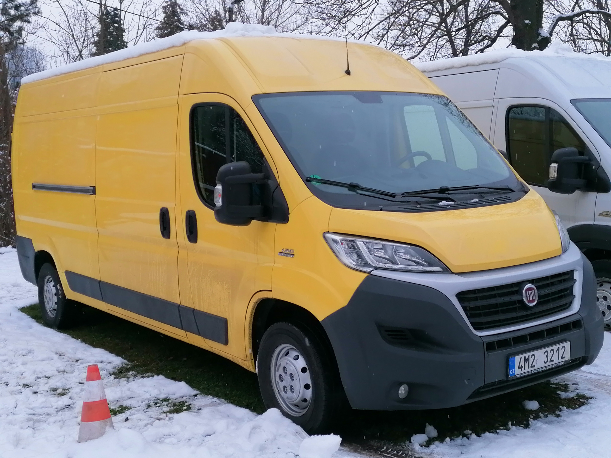 Żółty furgon dostawczy marki Fiat Ducato zaparkowany na śniegu, z pomarańczowo-białym pachołkiem drogowym w pobliżu przedniego koła.