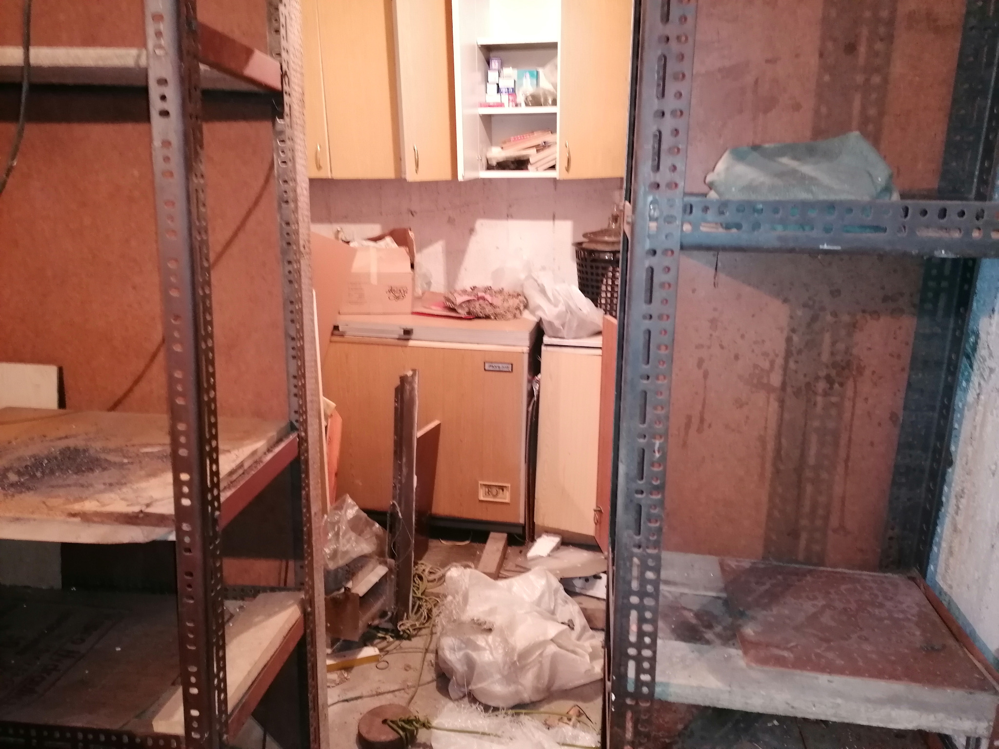 Metalowe regały magazynowe z różnymi przedmiotami, w tym workami, pudłami i sprzętem AGD, w pomieszczeniu o nierównym oświetleniu i widocznych śladach użytkowania.