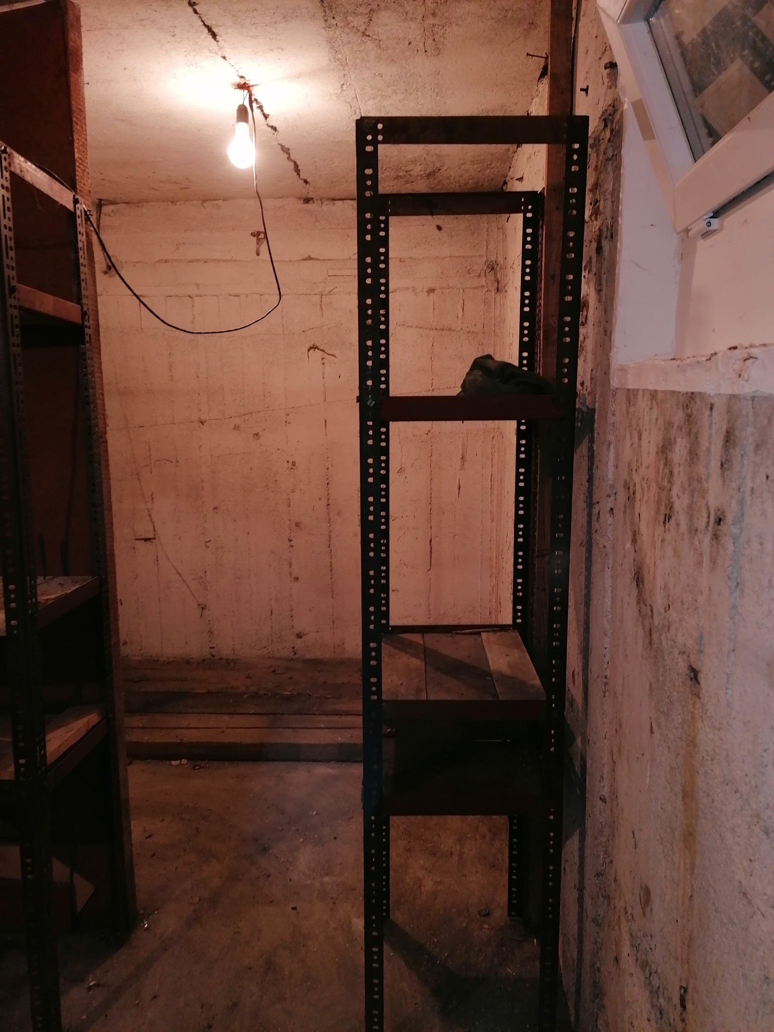 Metalowy regał z drewnianymi półkami w piwnicy, pod żarówką, z wilgocią na ścianach.