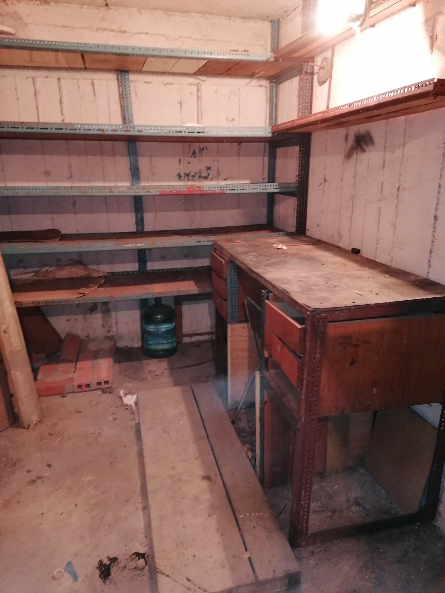Niedostatecznie oświetlone pomieszczenie gospodarcze z metalowymi regałami, drewnianym blatem roboczym z otwartą szufladą, cegłami i butelką wody mineralnej na podłodze.