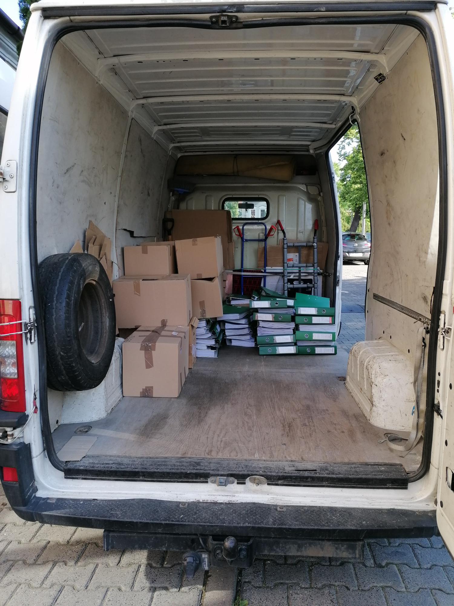 Wnętrze białego busa załadowane kartonami, stosem segregatorów z dokumentami i kołem zapasowym, widok z zewnątrz przez otwarte tylne drzwi.