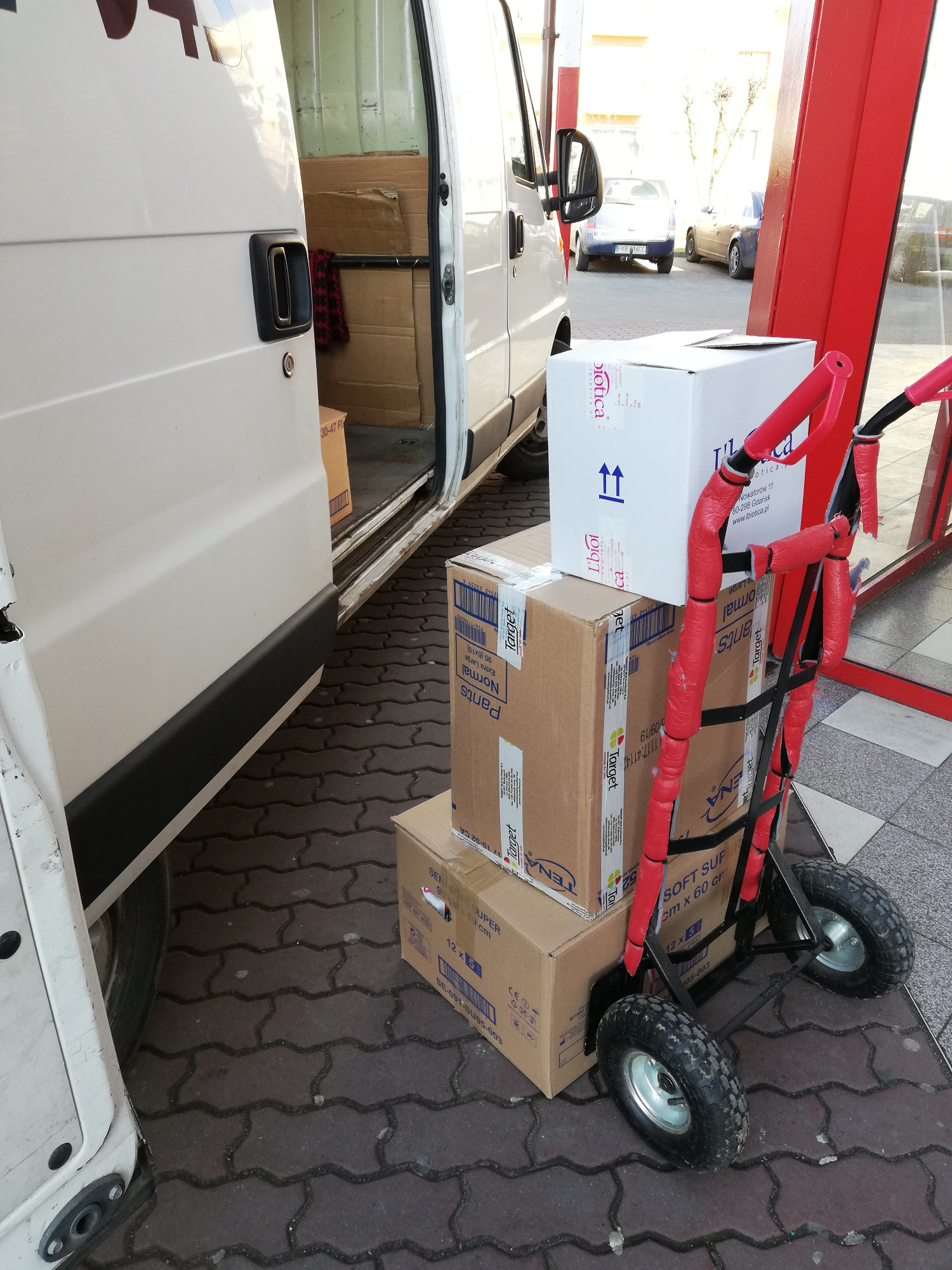 Wózek transportowy z trzema kartonami przed otwartym busem dostawczym na chodniku z kostki brukowej.