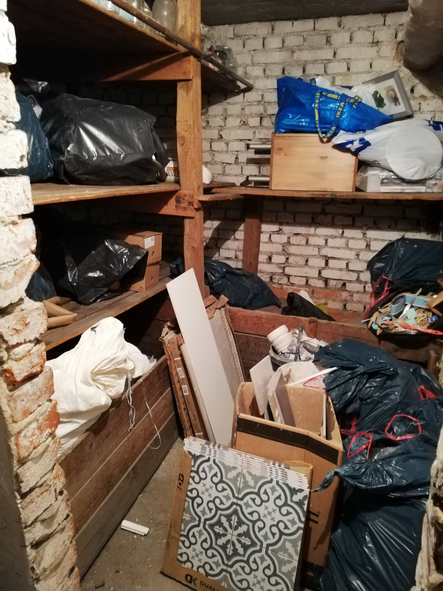 Zagracony stary strych z ceglanymi ścianami i drewnianymi półkami, na których znajdują się czarne worki na śmieci, kartony i płytki ceramiczne w pudełku.