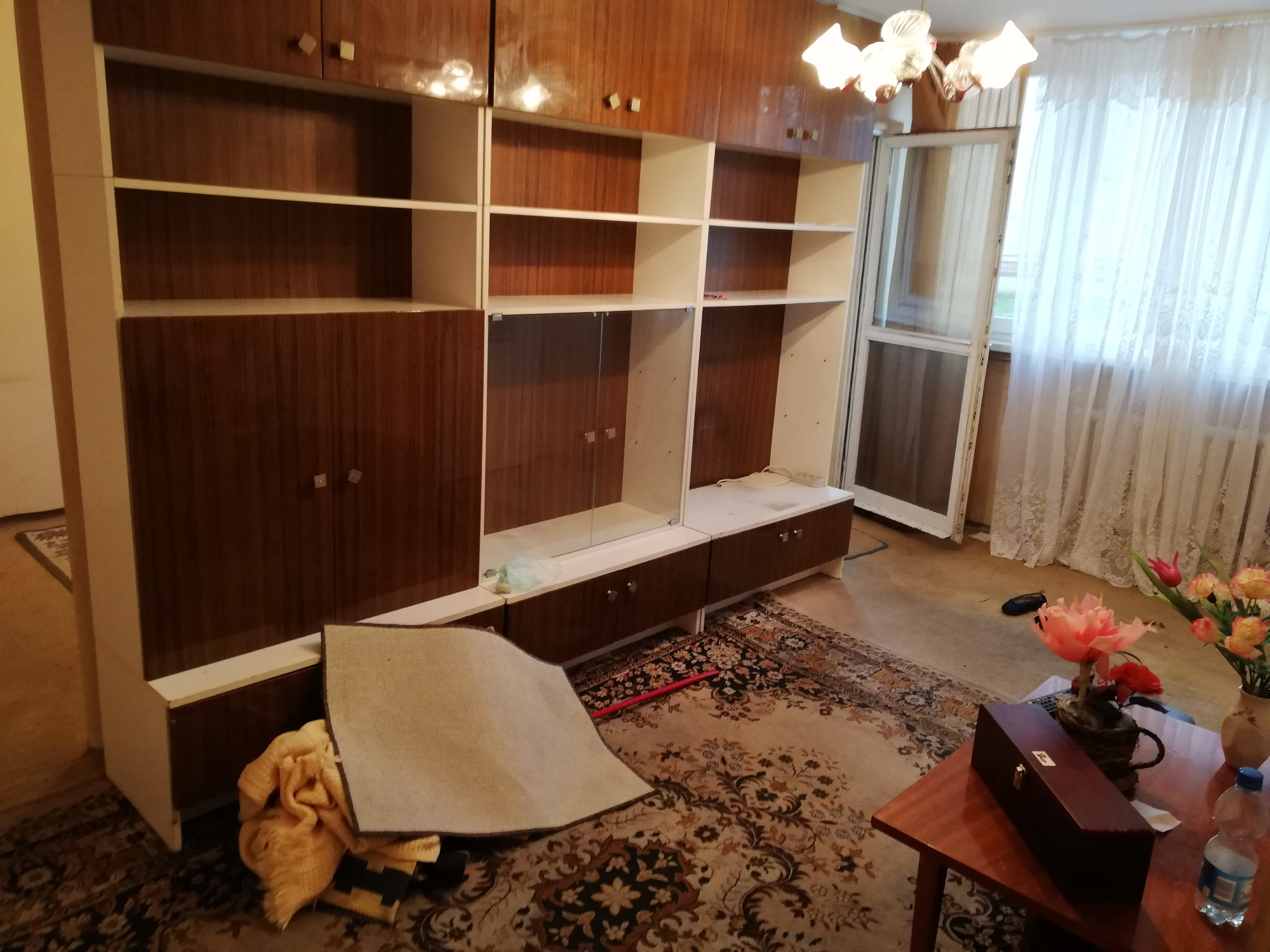 Demontaż meblościanki w trakcie opróżniania mieszkania, widoczny dywan na podłodze i kwiaty na stoliku w tle.