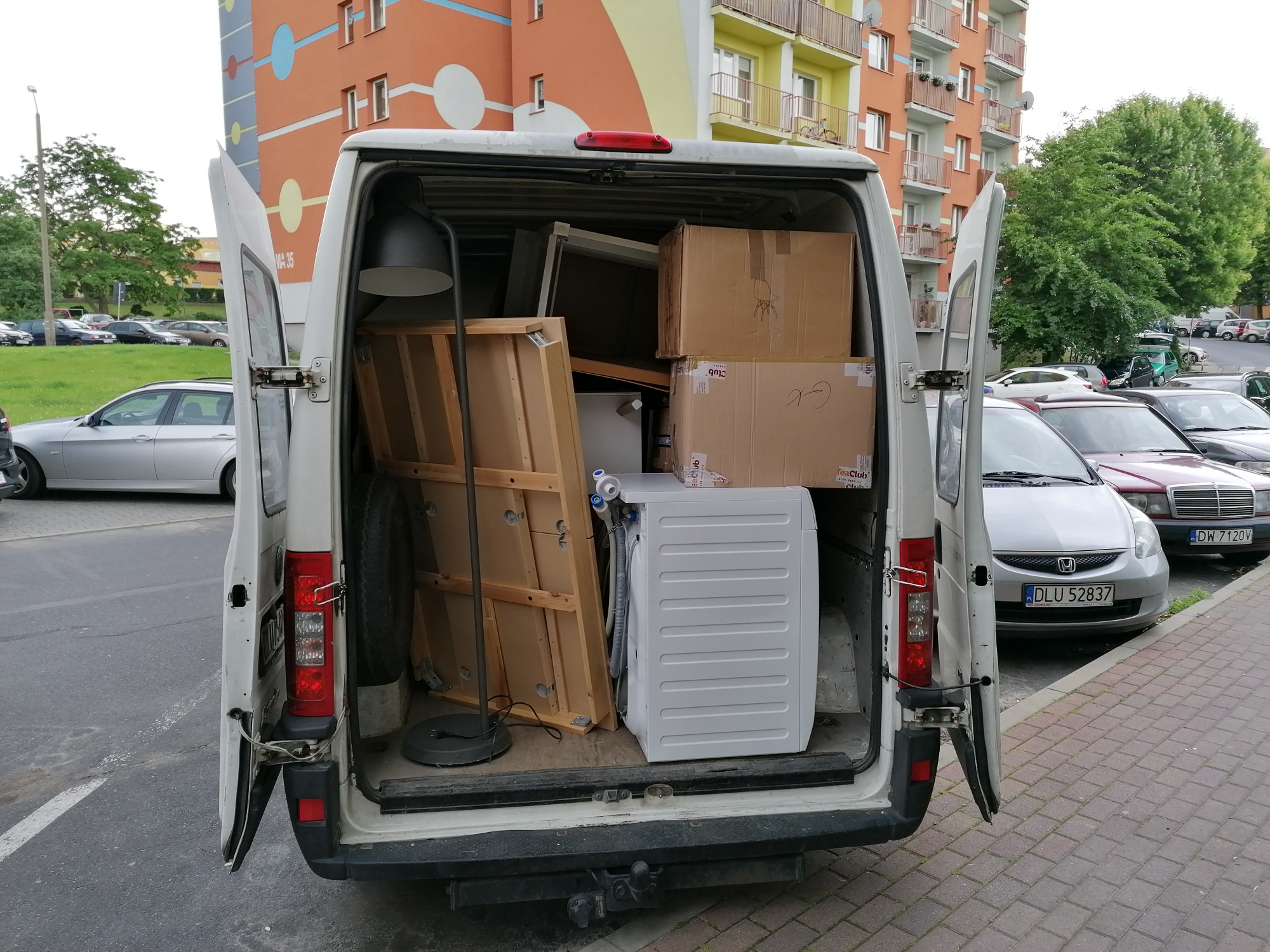 Otwarty furgon wypełniony sprzętem AGD, meblami i kartonami podczas przeprowadzki w Lubinie, widok z tyłu na chodniku z zaparkowanymi samochodami i blokiem mieszkalnym w tle.