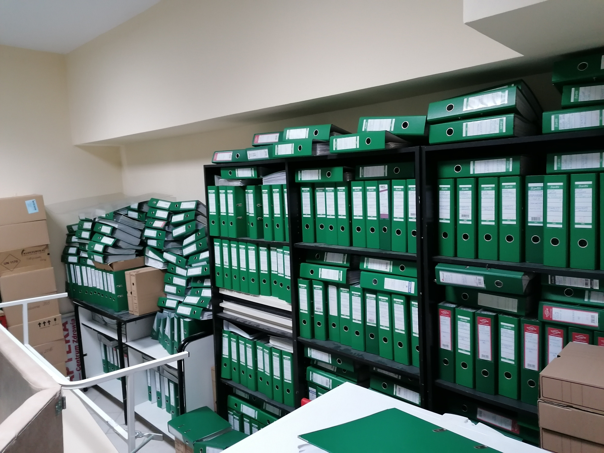 Archiwum biurowe: regały wypełnione rzędami zielonych segregatorów, część z nich ułożona w stosy na półkach i na wózku transportowym, obok widoczne kartonowe pudła z oznaczeniami ostrzegawczymi.