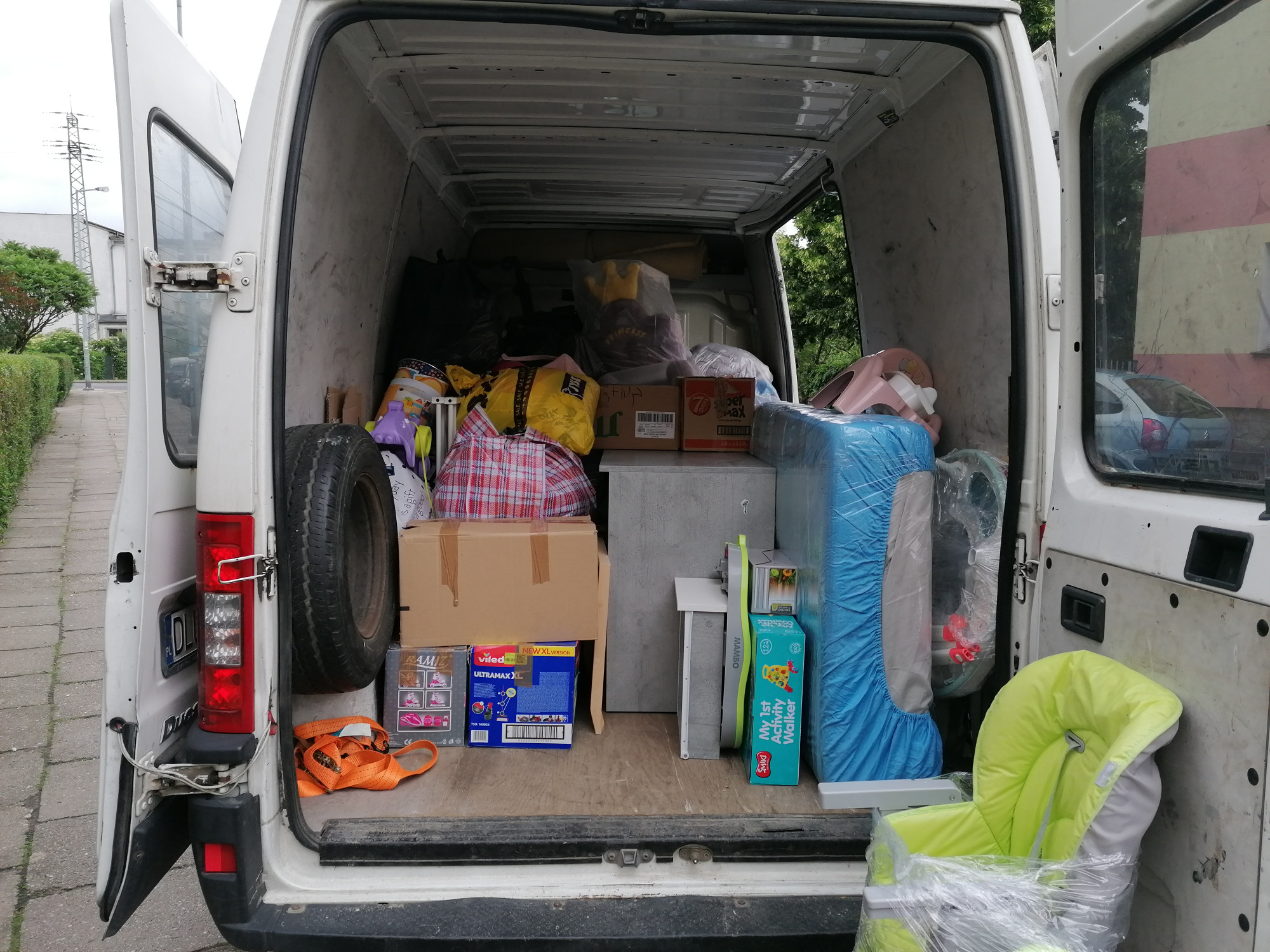 Wnętrze białego busa z otwartymi drzwiami załadowane różnorodnymi przedmiotami do transportu, w tym kartonami, workami i fotelikiem dziecięcym owiniętym folią. Widoczne opony zapasowe i pasy...