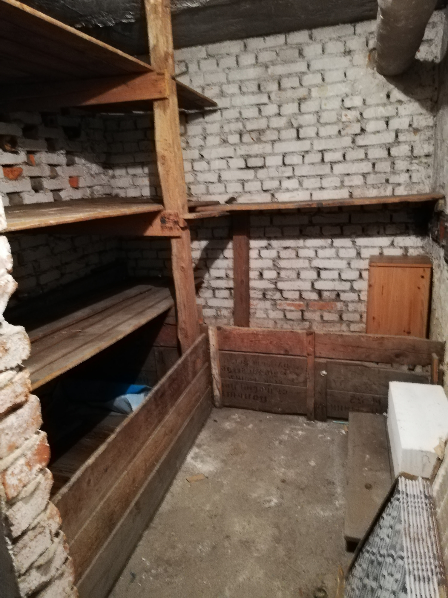 Piwnica z drewnianymi półkami i skrzyniami, ściany z białej cegły, surowy beton na podłodze, widoczne elementy konstrukcyjne i rury pod sufitem.