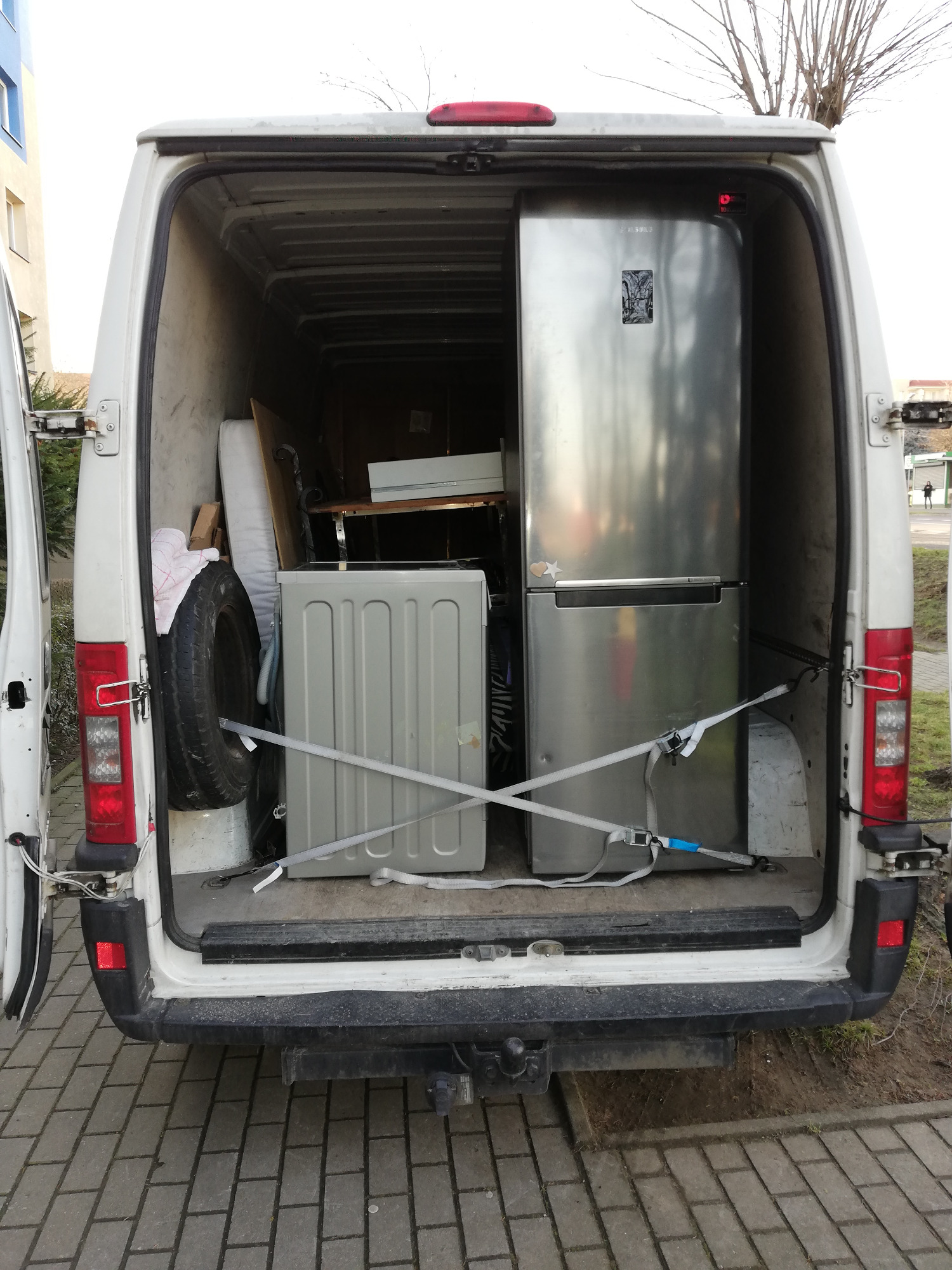 Wnętrze białego busa z otwartymi drzwiami, załadowane sprzętem AGD: srebrną lodówką Samsung i pralką, zabezpieczonymi pasami transportowymi, widoczne również opona i inne przedmioty.
