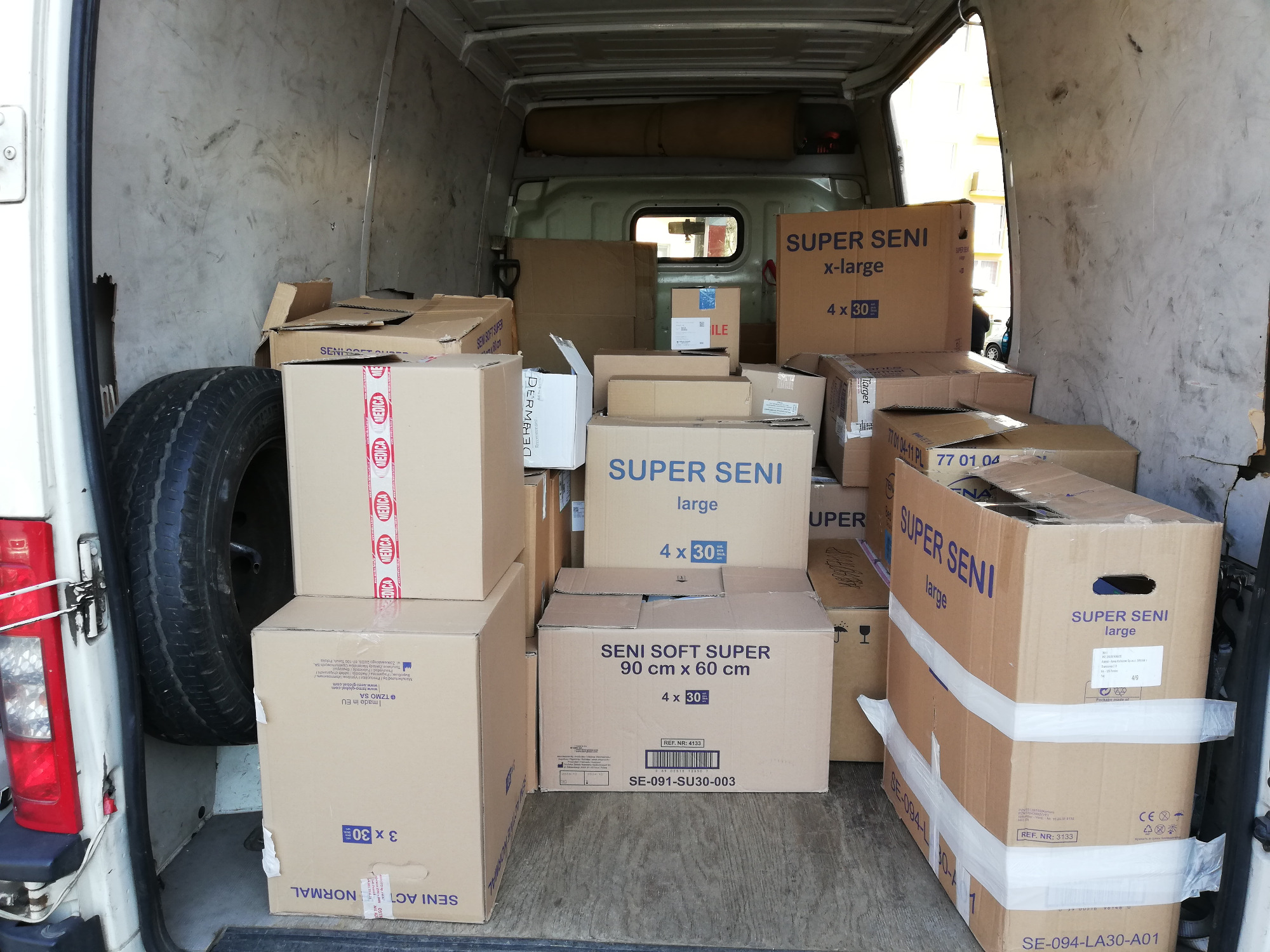 Wnętrze furgonetki wypełnione kartonami z produktami higienicznymi marki Seni, widoczne zapasowe koło, oświetlenie dzienne z zewnątrz.