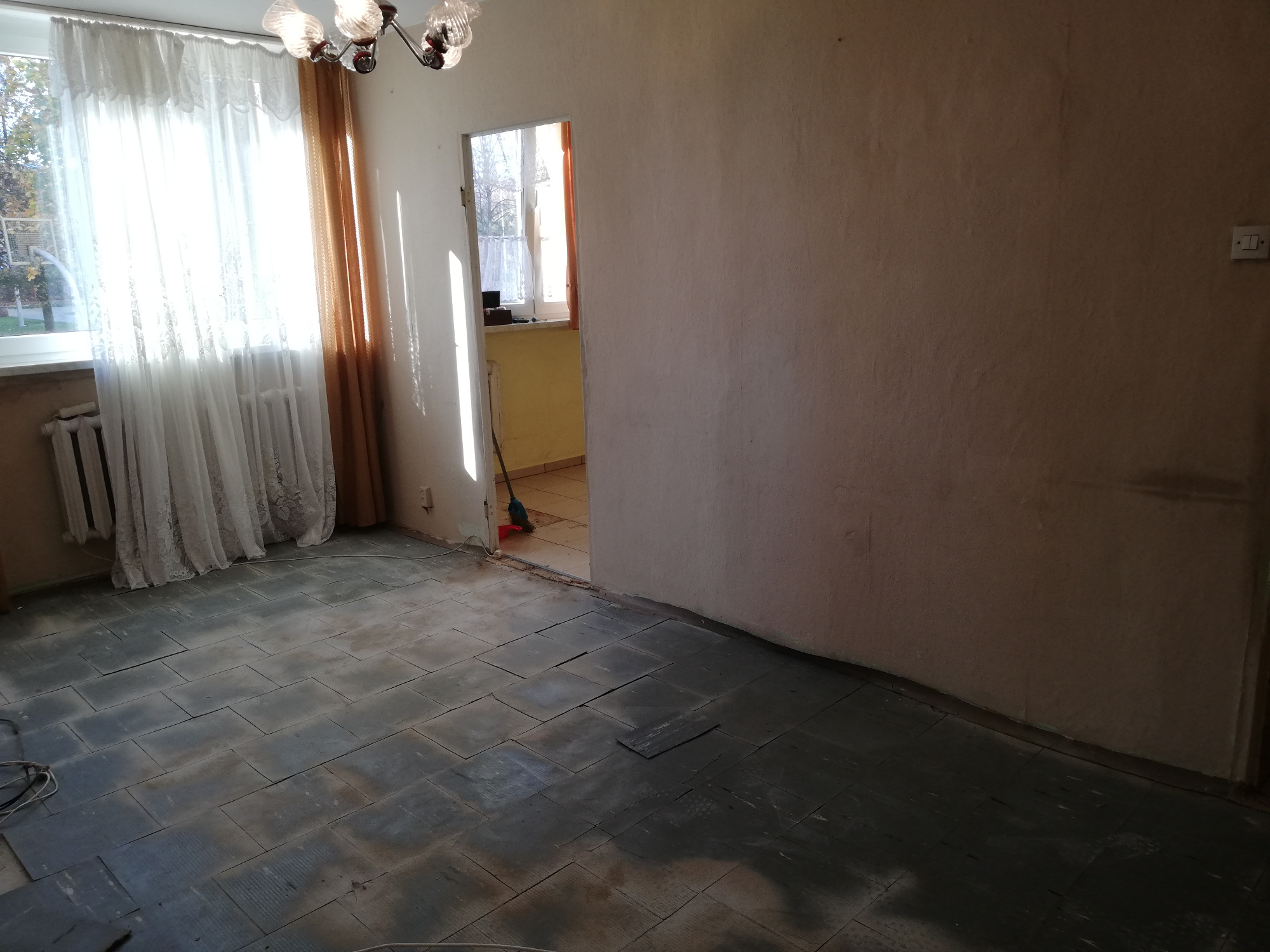 Pokój w trakcie demontażu podłogi z płytek, widoczne ślady po kleju i częściowo usunięte płytki, ściany w stanie surowym, w tle widoczne okno z firanką.