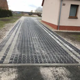 Długa, nowo ułożona droga z szaro-czarnej kostki brukowej typu I, prowadząca w kierunku domu z numerem 20, otoczona trawnikiem i tujami.