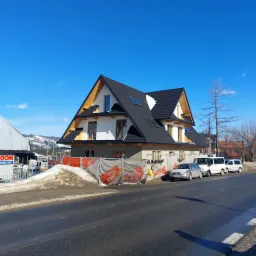 Nowoczesny dom szkieletowy w trakcie budowy, widok z zewnątrz, czarny dach z oknami dachowymi, biała elewacja, widoczne rusztowania i zabezpieczenia budowy, w tle góry.