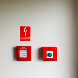 IN-TECH PIOTR KAPUŚCIŃSKI - Przeciwpożarowy wyłącznik prądu oraz przycisk uruchamiający PWP zamontowane na ścianie w Wieliszewie, obok naklejka ostrzegawcza o wysokim napięciu.