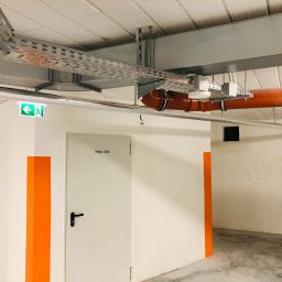 IN-TECH PIOTR KAPUŚCIŃSKI - Metalowe korytko kablowe pod sufitem, pomarańczowa rura, białe ściany z pomarańczowymi pasami, drzwi z napisem POM-GOS i zielony znak ewakuacyjny.