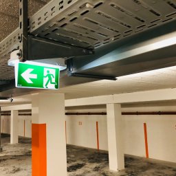 IN-TECH PIOTR KAPUŚCIŃSKI - Oznakowanie drogi ewakuacyjnej w garażu podziemnym, widoczny zielony piktogram z podświetleniem LED, metalowe koryta kablowe pod sufitem.