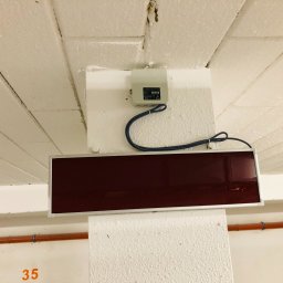 IN-TECH PIOTR KAPUŚCIŃSKI - Czujnik dymu z panelem informacyjnym zamontowany na suficie w garażu podziemnym, widoczne rury instalacyjne i numer stanowiska.