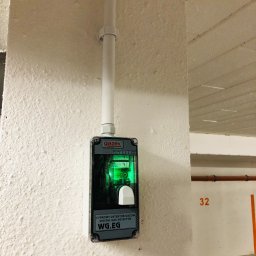 IN-TECH PIOTR KAPUŚCIŃSKI - Cyfrowy detektor gazów WG.EG marki Gazex zamontowany na pomalowanej na biało ścianie w garażu podziemnym, z widoczną rurką instalacyjną powyżej.