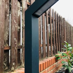 IN-TECH PIOTR KAPUŚCIŃSKI - Nowoczesna lampa ogrodowa w kolorze grafitowym, oświetlająca drewniany płot z ceglaną podmurówką i młodymi roślinami.