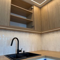 IN-TECH PIOTR KAPUŚCIŃSKI - Kuchnia z drewnianymi szafkami, czarnym zlewem i baterią. Oświetlenie LED podszafkowe i sufitowe. Otwarta szafka eksponuje półki.