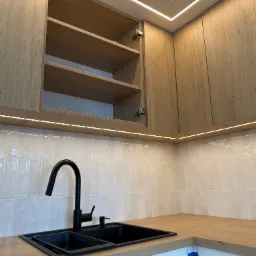 Kuchnia z drewnianymi szafkami, czarnym zlewem i baterią. Oświetlenie LED podszafkowe i sufitowe. Otwarta szafka eksponuje półki.