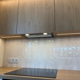 IN-TECH PIOTR KAPUŚCIŃSKI - Kuchnia z drewnianymi szafkami, okapem i oświetleniem LED pod szafkami. Płyta indukcyjna na blacie. Białe, błyszczące płytki na ścianie i gniazdka elektryczne.