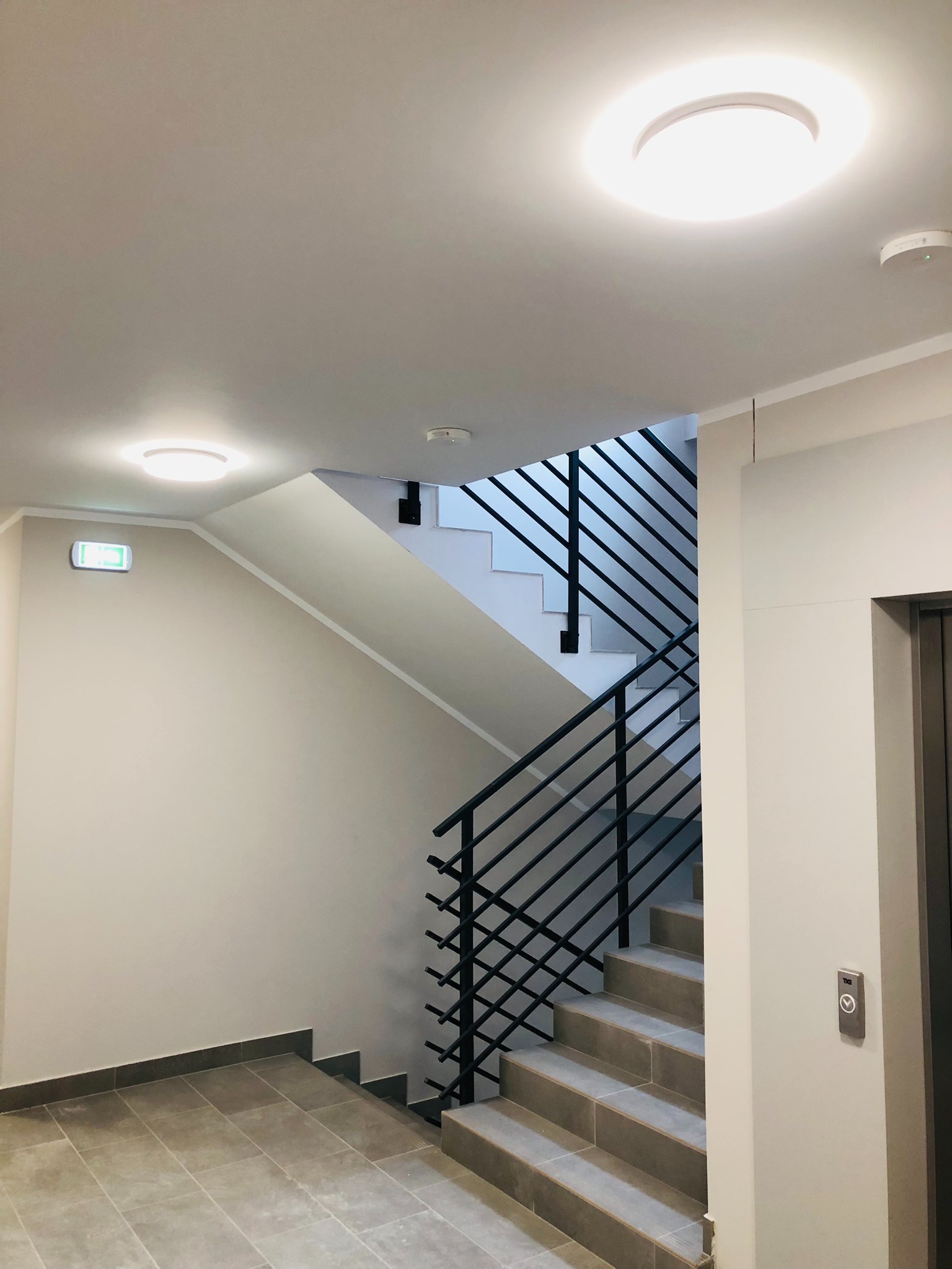 Klatka schodowa w nowoczesnym budynku z szarymi płytkami na podłodze, oświetleniem sufitowym i czarną balustradą. Widoczny również przycisk windy oraz znak ewakuacyjny.