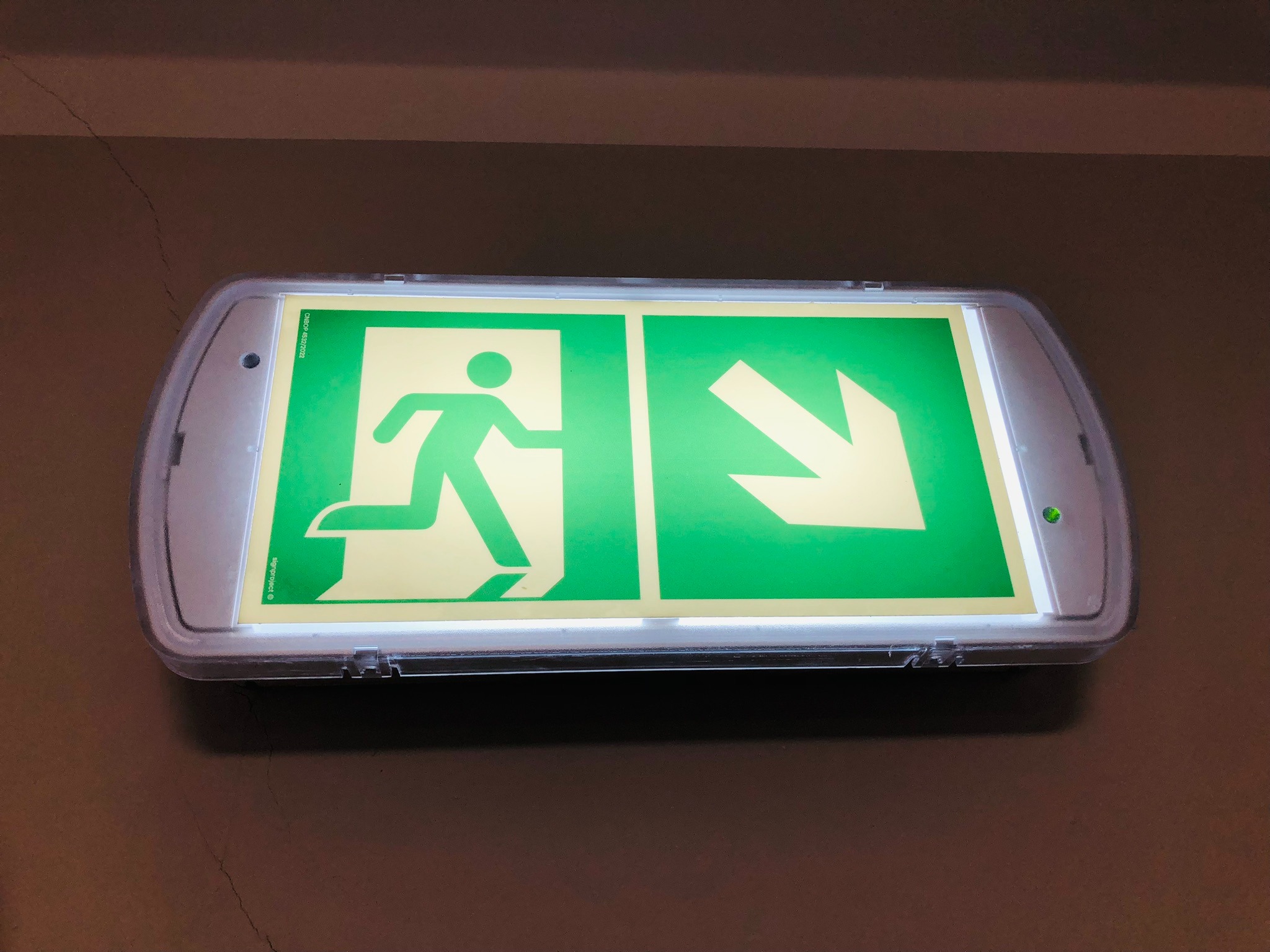 Podświetlany, biało-zielony znak ewakuacyjny z symbolem biegnącej osoby i strzałką w dół, zamontowany na brązowej ścianie.