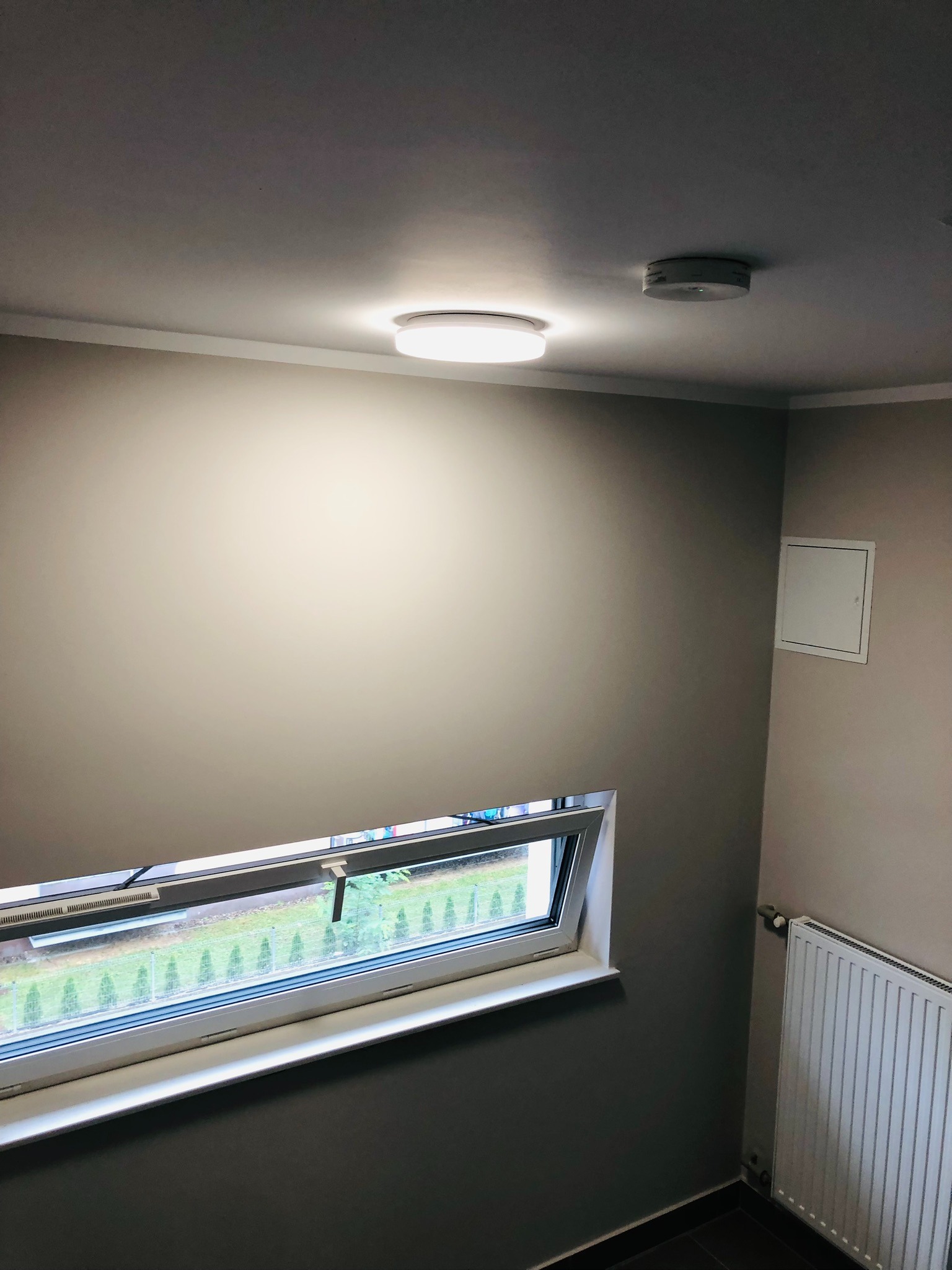 Nowoczesne, okrągłe oświetlenie sufitowe LED, czujnik dymu, okno z widokiem na ogród i grzejnik w szarej klatce schodowej.