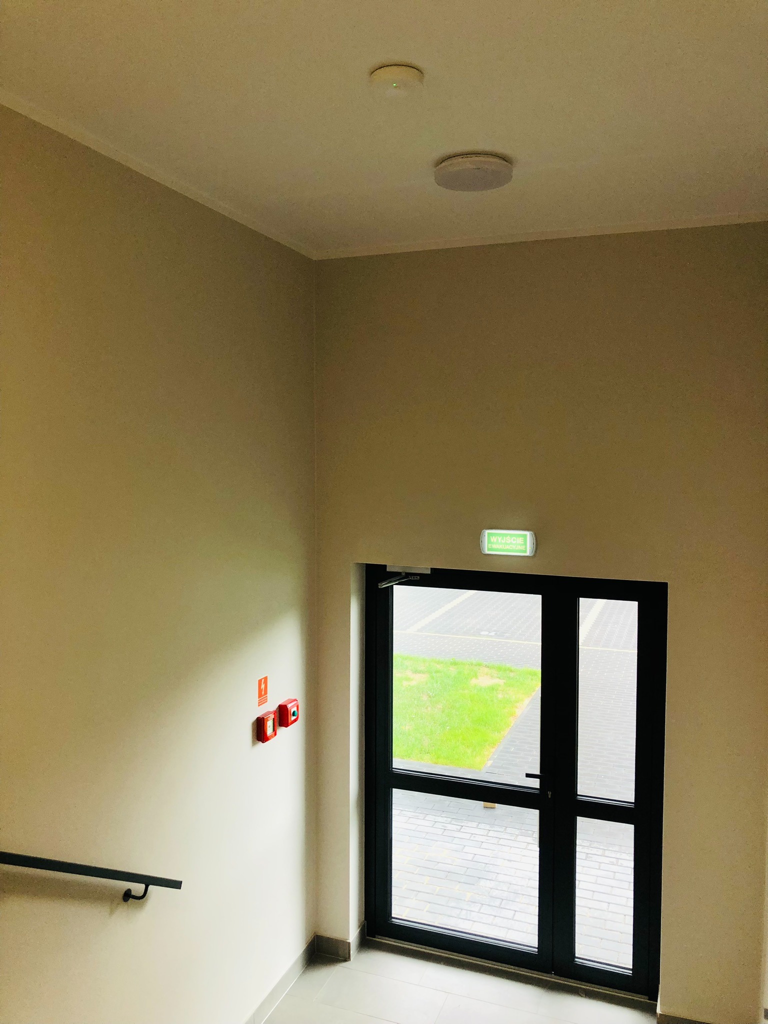 Klatka schodowa z czarną ramą drzwi ewakuacyjnych, zielony znak 'Wyjście Ewakuacyjne', czerwone przyciski alarmowe i poręcz schodowa.