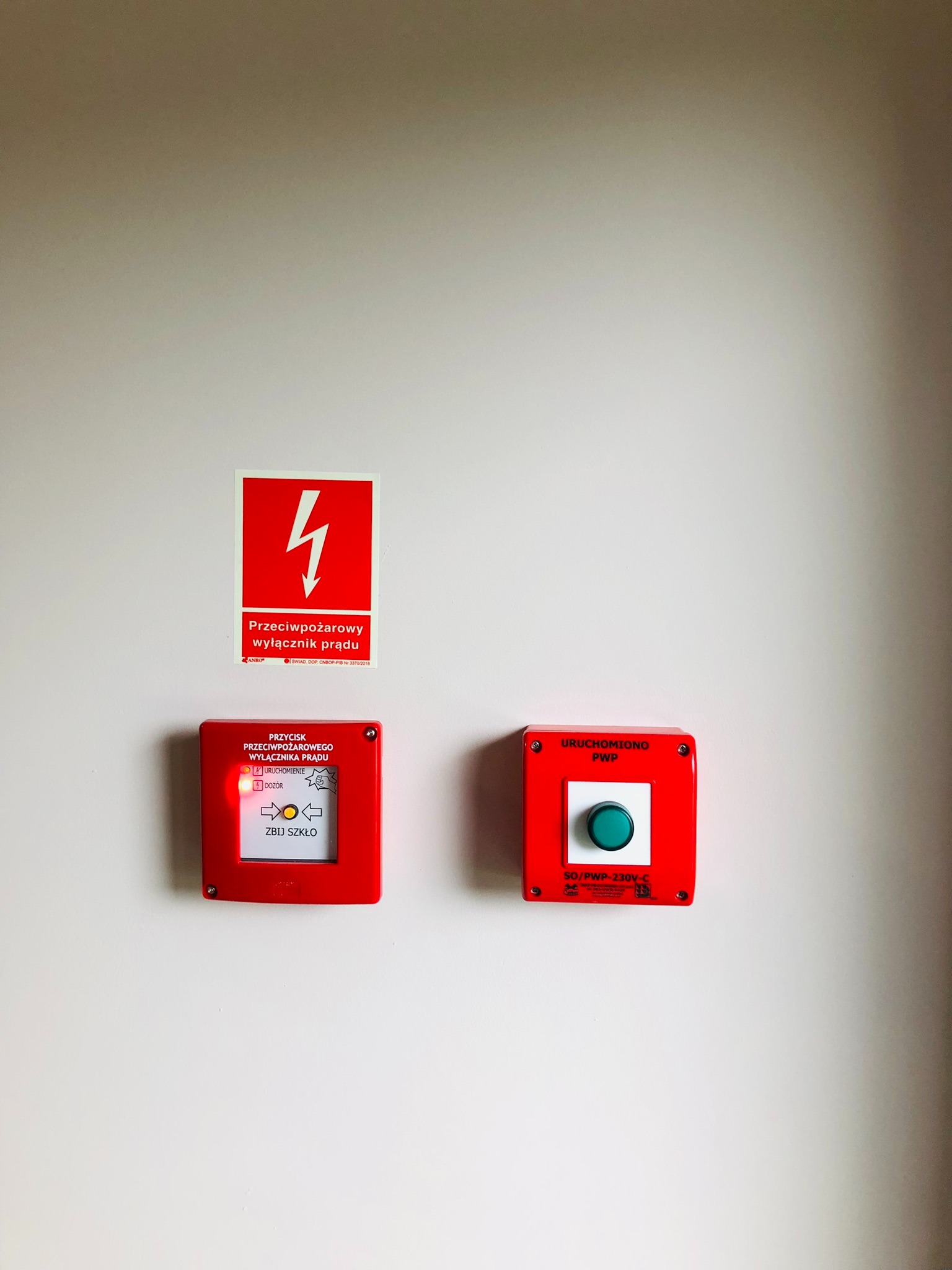 Przeciwpożarowy wyłącznik prądu oraz przycisk uruchamiający PWP zamontowane na ścianie w Wieliszewie, obok naklejka ostrzegawcza o wysokim napięciu.