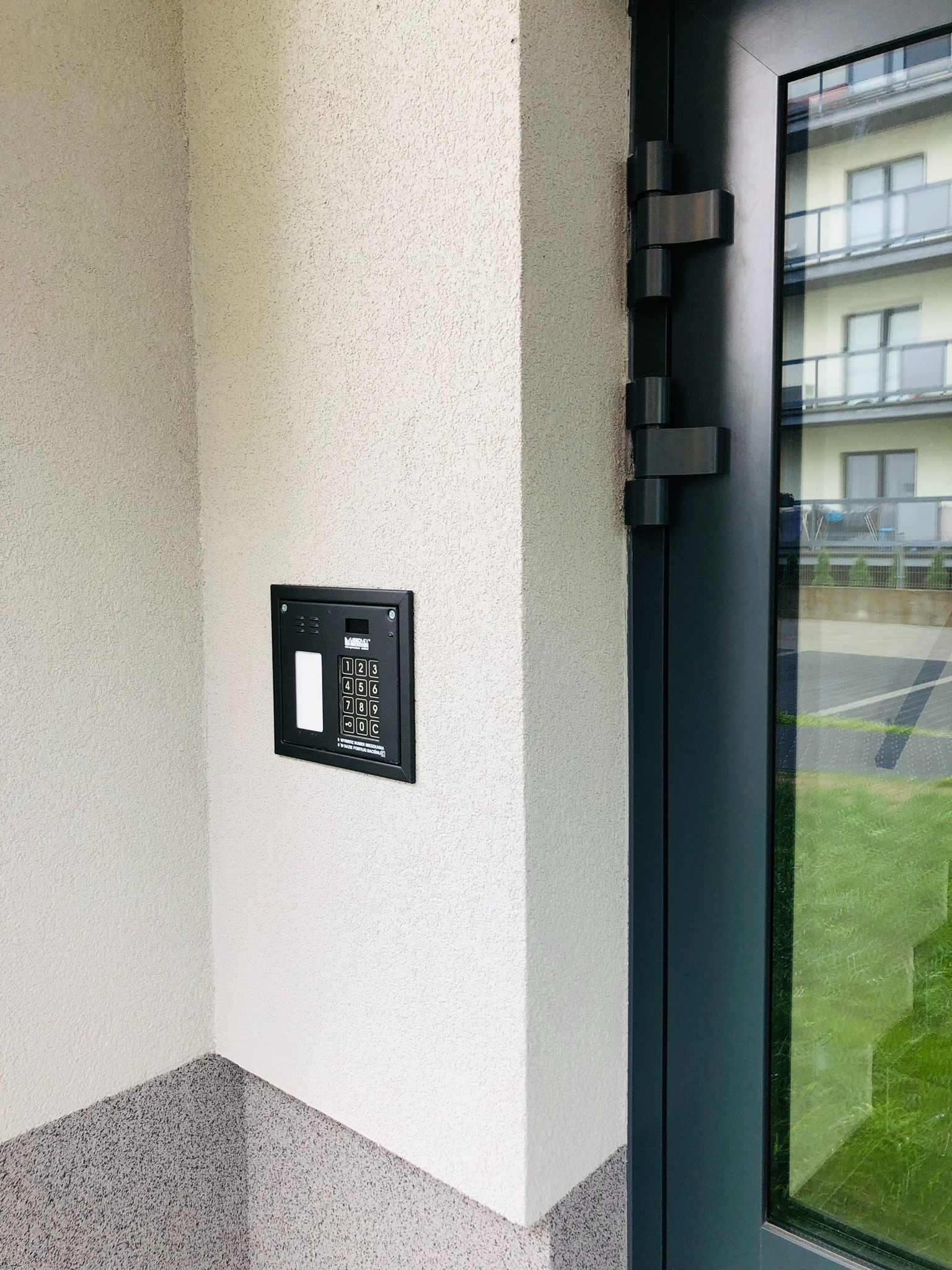 Zewnętrzny panel domofonu z klawiaturą numeryczną zamontowany na elewacji budynku przy wejściu.