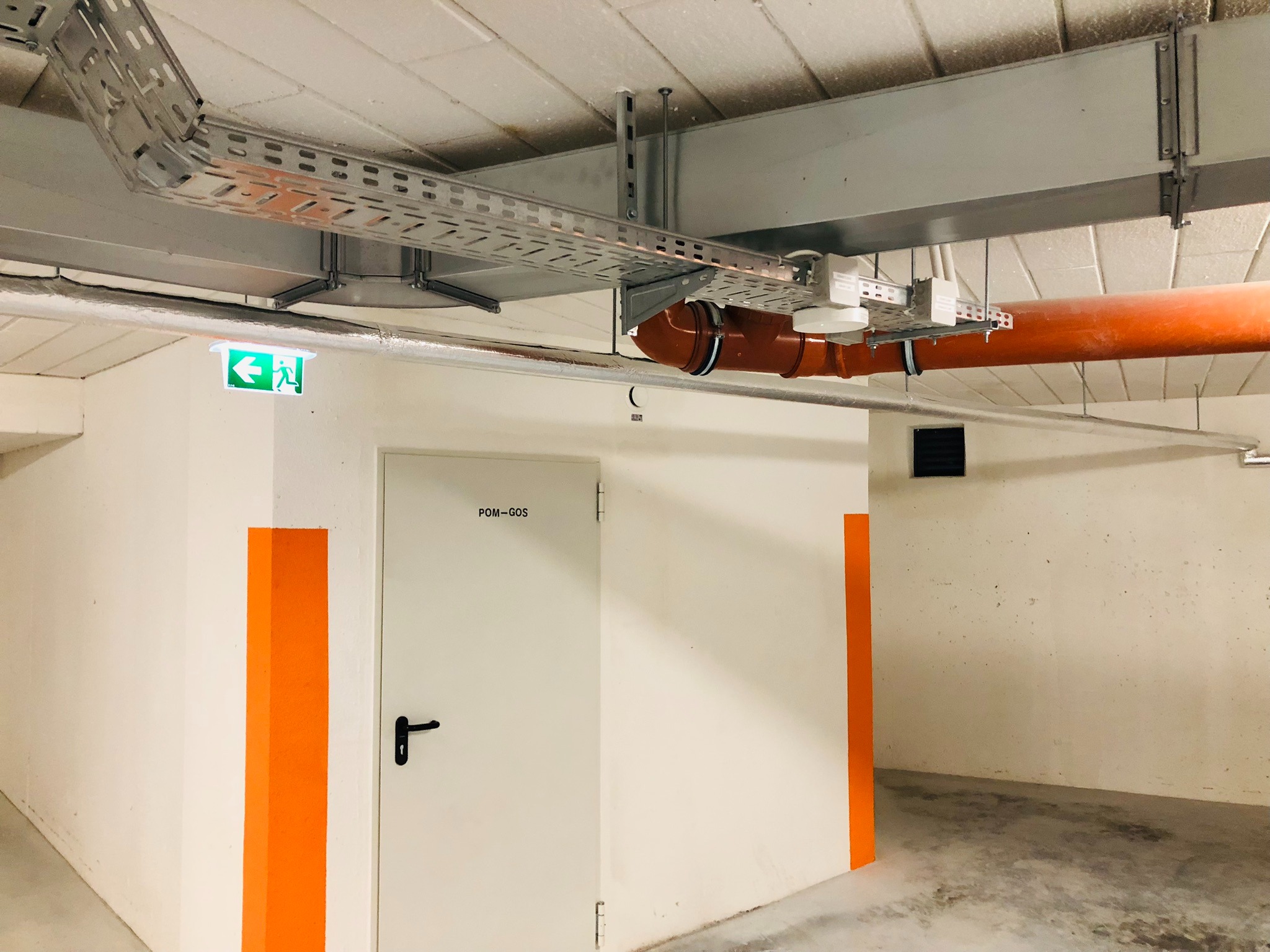 Metalowe korytko kablowe pod sufitem, pomarańczowa rura, białe ściany z pomarańczowymi pasami, drzwi z napisem POM-GOS i zielony znak ewakuacyjny.
