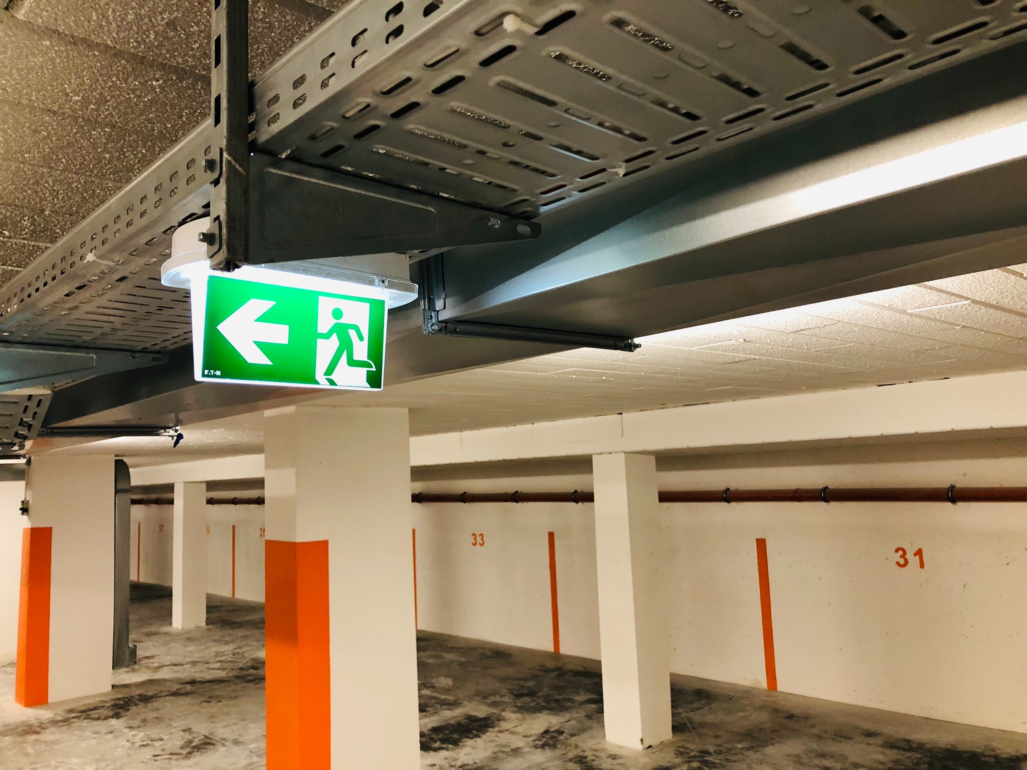 Oznakowanie drogi ewakuacyjnej w garażu podziemnym, widoczny zielony piktogram z podświetleniem LED, metalowe koryta kablowe pod sufitem.