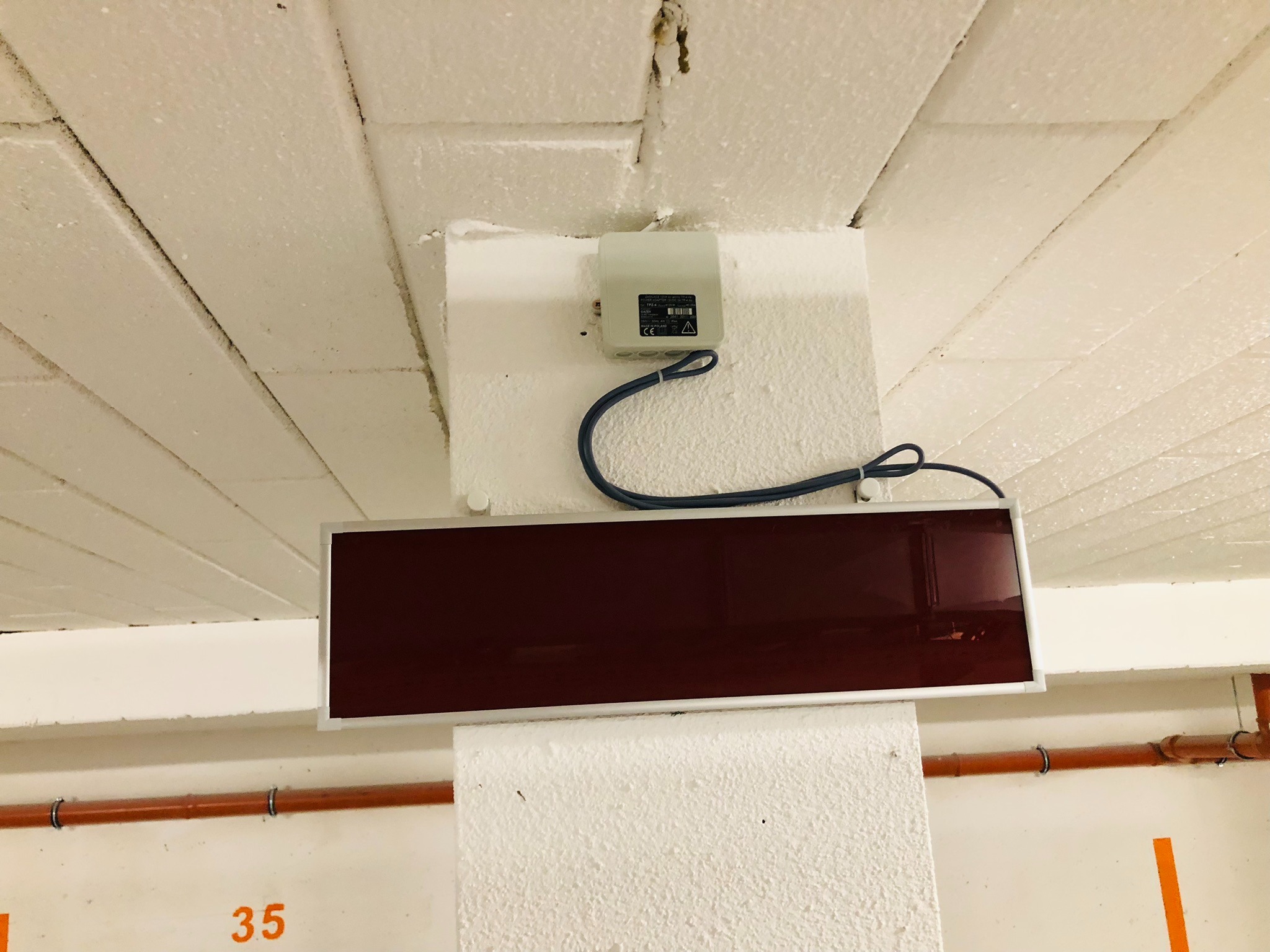 Czujnik dymu z panelem informacyjnym zamontowany na suficie w garażu podziemnym, widoczne rury instalacyjne i numer stanowiska.